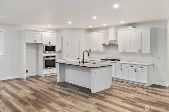 kitchen featuring wall chimney range hood, a sink, light wood-type flooring, stainless steel appliances, and white cabinets