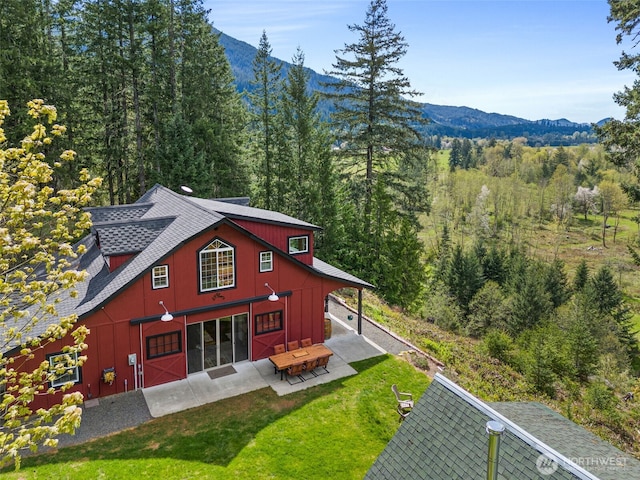 back of property with a mountain view, a shingled roof, a patio area, and a wooded view