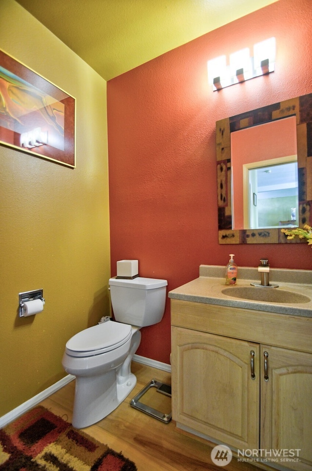 half bath with toilet, vanity, wood finished floors, and baseboards