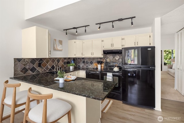 kitchen with black appliances, a sink, a peninsula, light wood-type flooring, and dark stone counters
