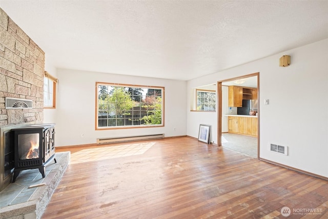 unfurnished living room featuring a wood stove, light wood finished floors, visible vents, baseboards, and a baseboard radiator