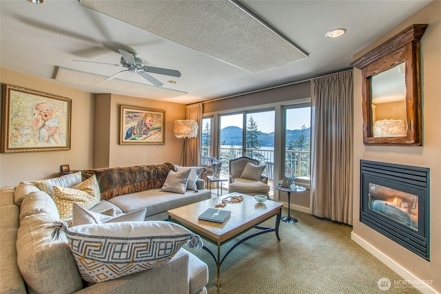 living room with a mountain view, carpet, a ceiling fan, baseboards, and a glass covered fireplace