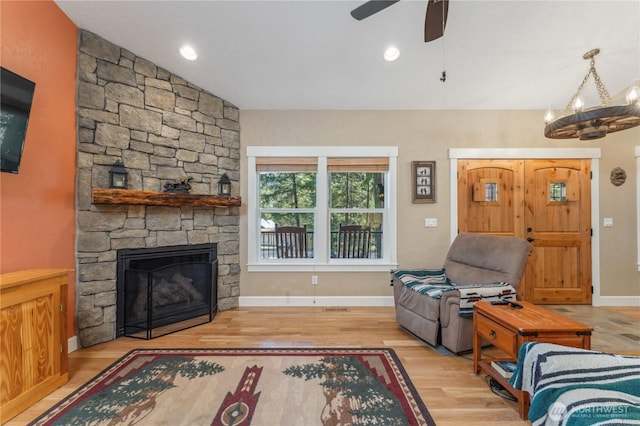 living area featuring baseboards, a stone fireplace, wood finished floors, and ceiling fan with notable chandelier