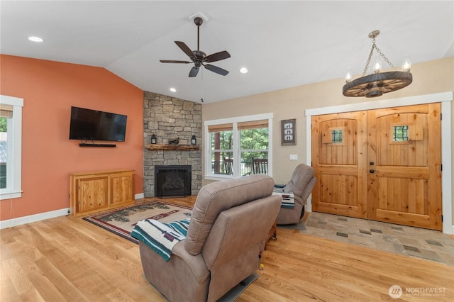 living area with wood finished floors, lofted ceiling, baseboards, a stone fireplace, and ceiling fan with notable chandelier