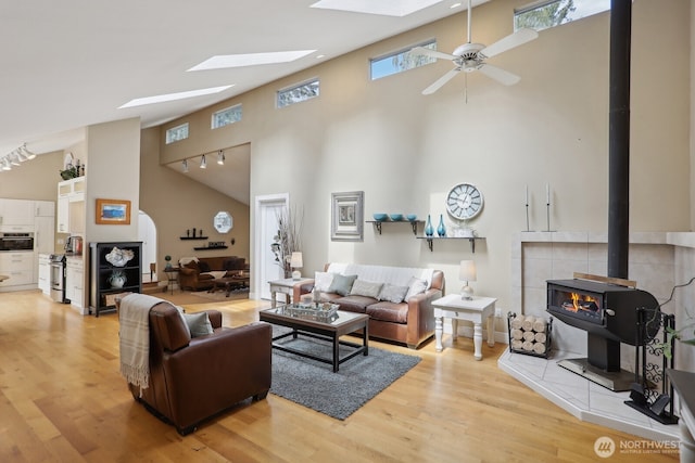 living area featuring high vaulted ceiling, a wood stove, a skylight, ceiling fan, and light wood finished floors