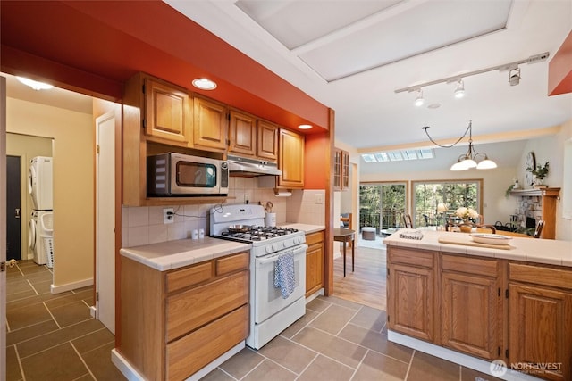 kitchen featuring stainless steel microwave, under cabinet range hood, white gas stove, tile countertops, and stacked washer / dryer