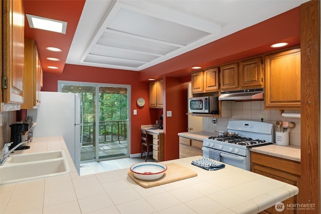 kitchen with a sink, under cabinet range hood, backsplash, tile countertops, and white appliances