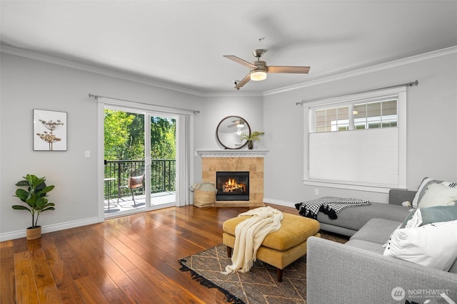 living area featuring crown molding, ceiling fan, wood-type flooring, baseboards, and a tile fireplace