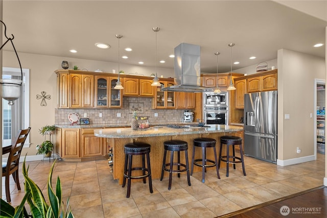 kitchen with light stone countertops, brown cabinets, stainless steel appliances, tasteful backsplash, and island range hood