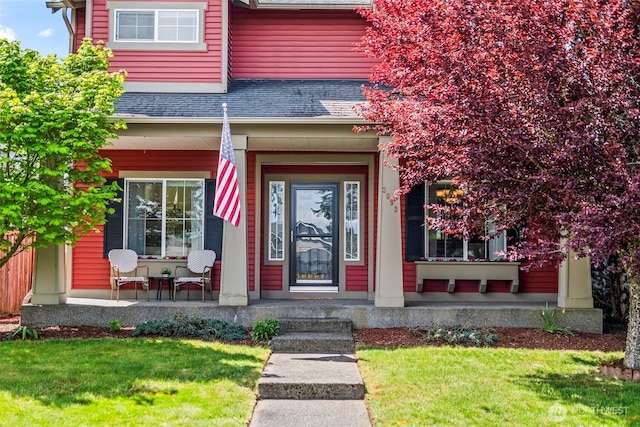 view of exterior entry with covered porch, a shingled roof, and a yard