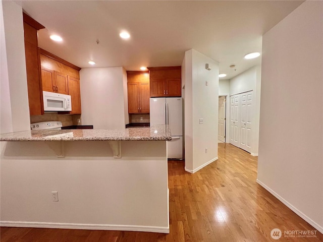 kitchen featuring brown cabinets, white appliances, light wood-style flooring, light stone counters, and a peninsula