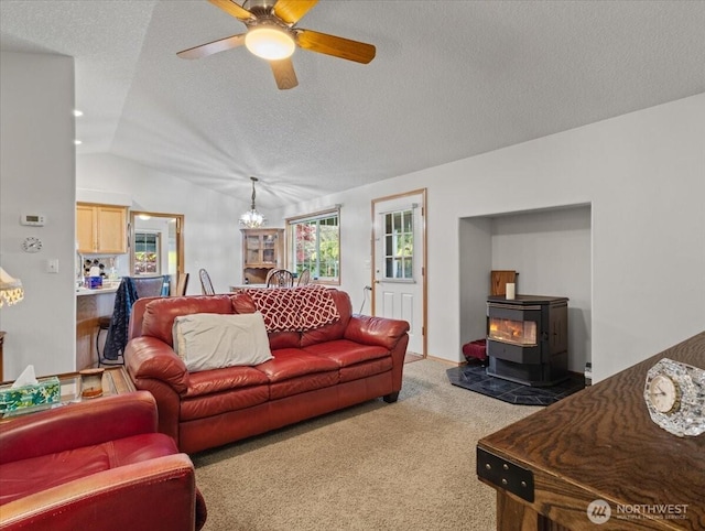 living area featuring a wood stove, vaulted ceiling, a textured ceiling, light colored carpet, and ceiling fan with notable chandelier