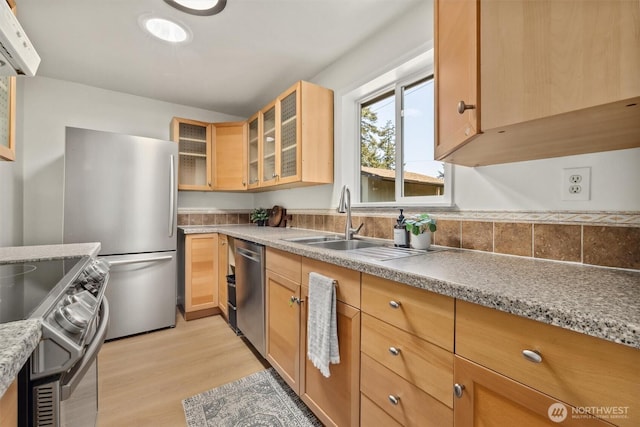 kitchen featuring appliances with stainless steel finishes, a sink, light wood finished floors, light brown cabinets, and glass insert cabinets