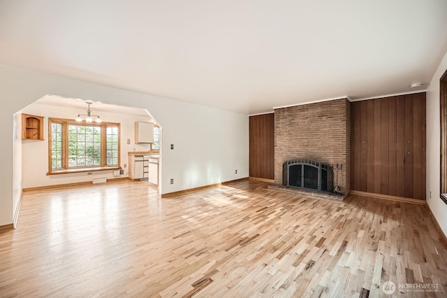 unfurnished living room featuring arched walkways, a brick fireplace, baseboards, light wood finished floors, and a chandelier