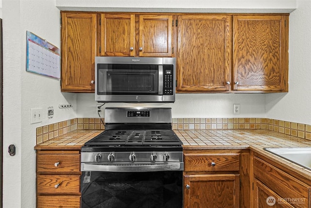 kitchen with brown cabinetry, tile countertops, a sink, and stainless steel appliances