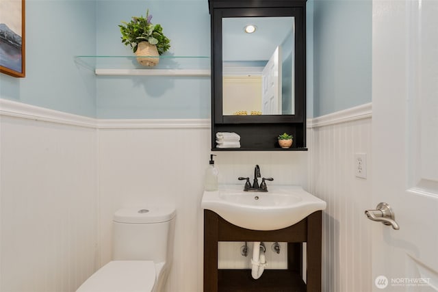 half bath with a sink, a decorative wall, a wainscoted wall, and toilet