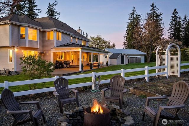 back of house at dusk with an outbuilding, fence, a storage unit, a yard, and a fire pit