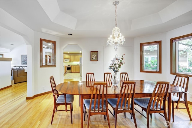 dining space with an inviting chandelier, a raised ceiling, light wood-style floors, arched walkways, and baseboards
