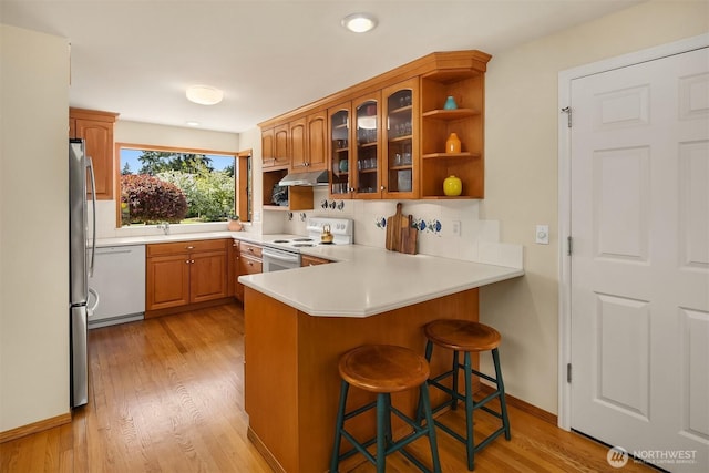 kitchen featuring a peninsula, white appliances, glass insert cabinets, under cabinet range hood, and brown cabinets
