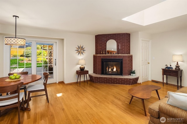 living area featuring a skylight, baseboards, a brick fireplace, and light wood-style flooring