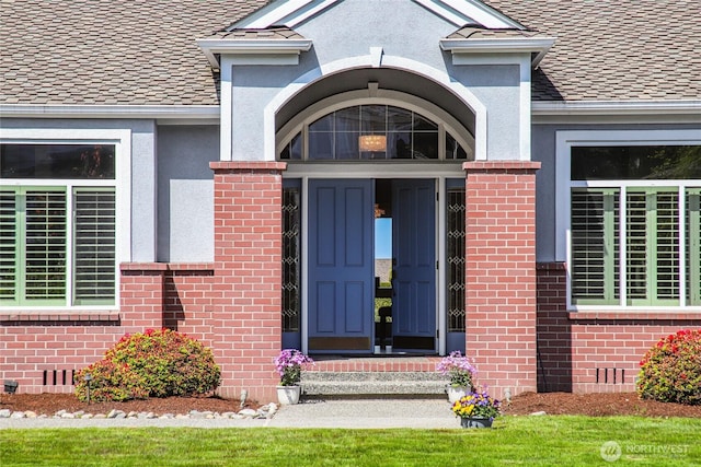 view of exterior entry featuring a shingled roof, stucco siding, and brick siding