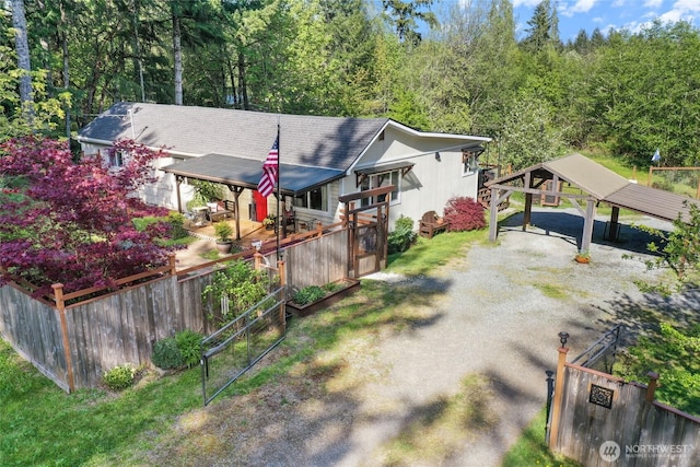 back of house featuring dirt driveway, a carport, a view of trees, roof with shingles, and fence