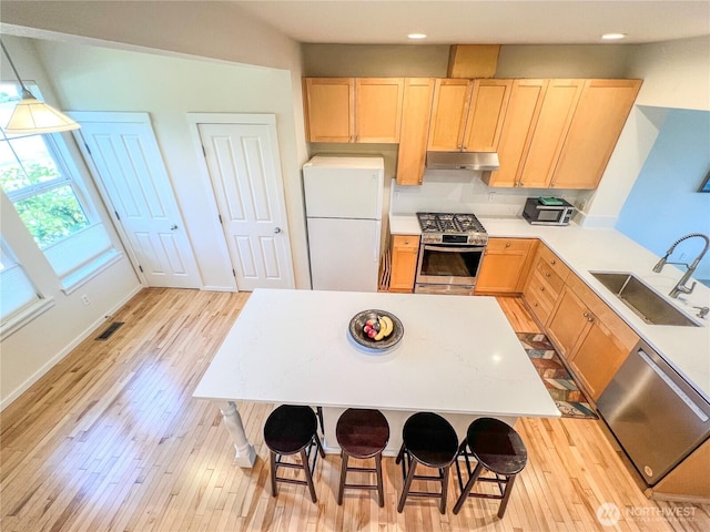 kitchen featuring light brown cabinetry, appliances with stainless steel finishes, a sink, light wood-style flooring, and under cabinet range hood