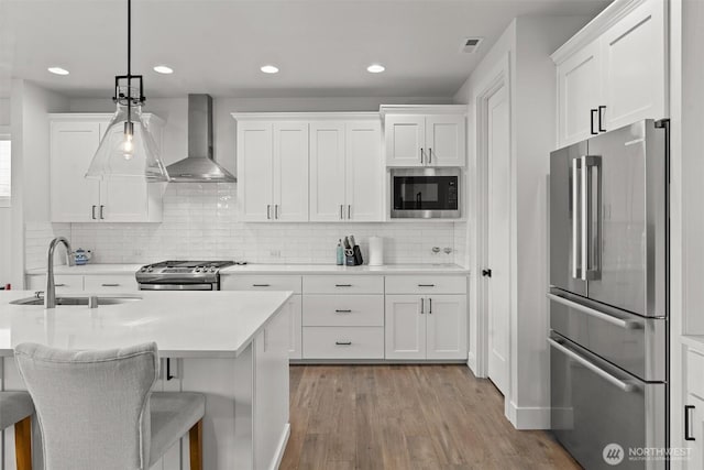 kitchen featuring wall chimney range hood, light wood-type flooring, light countertops, a sink, and appliances with stainless steel finishes