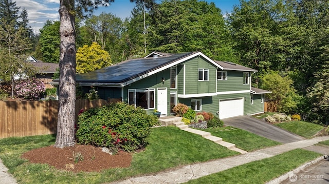 tri-level home featuring a front lawn, aphalt driveway, fence, an attached garage, and solar panels