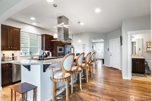 kitchen featuring island exhaust hood, dark brown cabinetry, visible vents, arched walkways, and appliances with stainless steel finishes