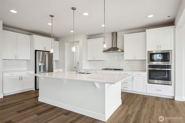 kitchen with wall chimney range hood, appliances with stainless steel finishes, a sink, a kitchen island with sink, and light countertops