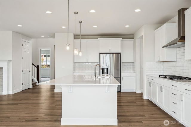 kitchen with wall chimney range hood, stainless steel appliances, a kitchen island with sink, and dark wood-style flooring