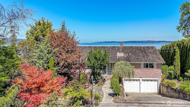 view of front of property featuring driveway, brick siding, a chimney, a garage, and a water view