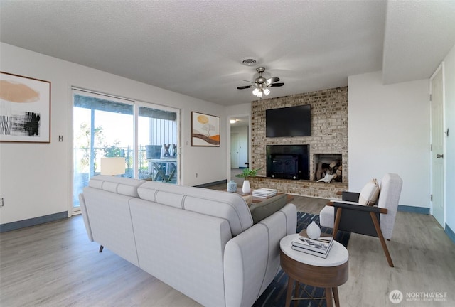 living room featuring light wood-style flooring, ceiling fan, baseboards, a textured ceiling, and a brick fireplace