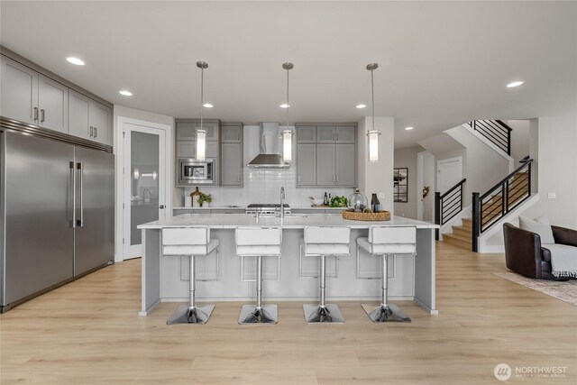 kitchen featuring wall chimney range hood, built in appliances, gray cabinets, light wood-type flooring, and a kitchen bar