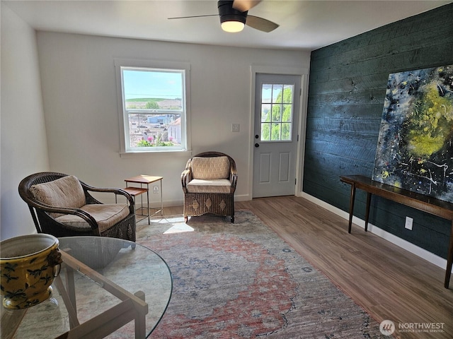 sitting room with wooden walls, a ceiling fan, baseboards, and wood finished floors