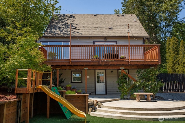 back of property featuring a playground, a wooden deck, a shingled roof, and fence