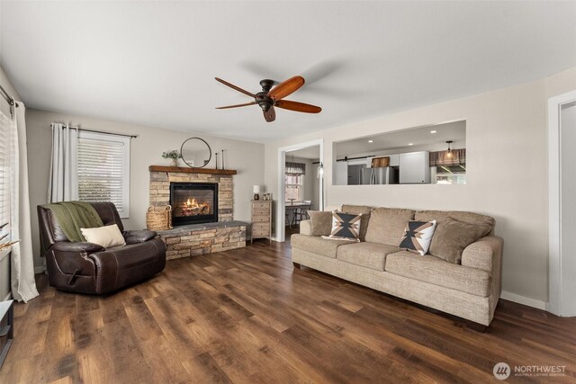 living area featuring a stone fireplace, a ceiling fan, baseboards, and dark wood-type flooring