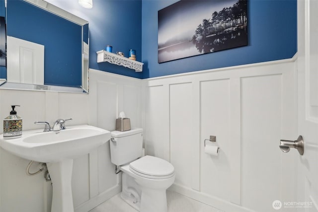 half bath featuring a decorative wall, toilet, wainscoting, a sink, and tile patterned flooring