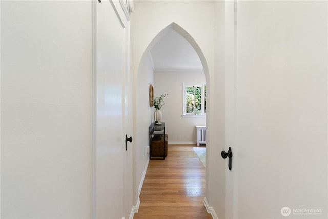 corridor featuring arched walkways, baseboards, radiator, and light wood-type flooring