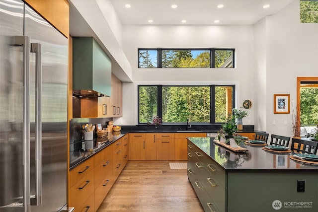 kitchen with a towering ceiling, a sink, wall chimney exhaust hood, high end refrigerator, and light wood-type flooring