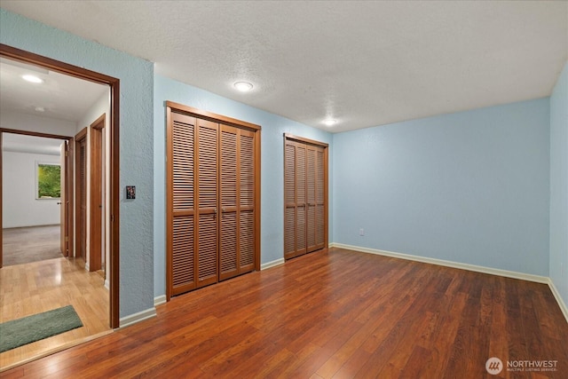 unfurnished bedroom with a textured wall, a textured ceiling, two closets, baseboards, and wood finished floors