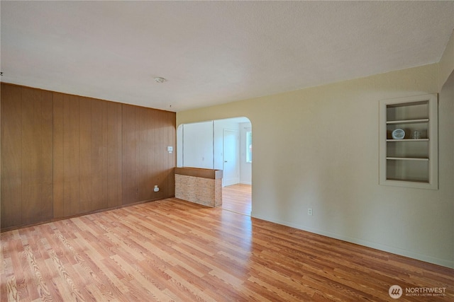 unfurnished room featuring light wood finished floors, built in shelves, arched walkways, baseboards, and wooden walls
