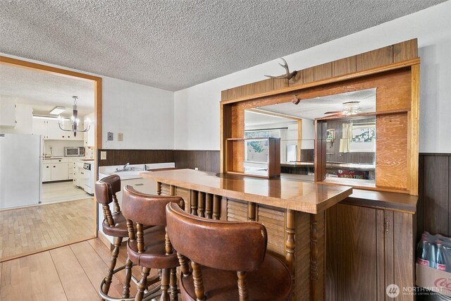 bar with light wood-style floors, freestanding refrigerator, a textured ceiling, and a wainscoted wall