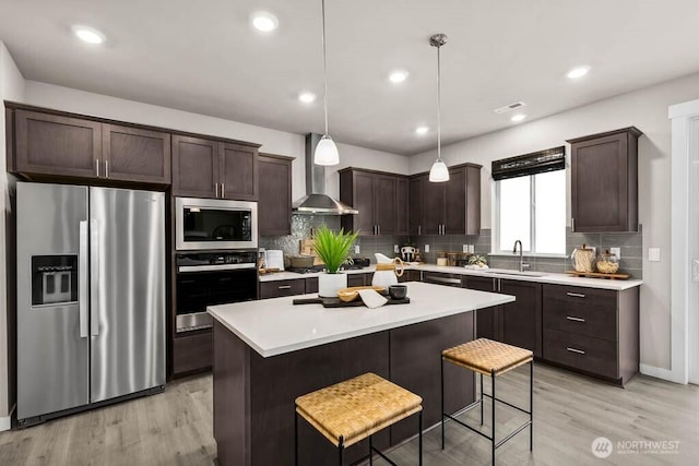 kitchen featuring wall chimney range hood, dark brown cabinets, light wood finished floors, stainless steel appliances, and decorative backsplash