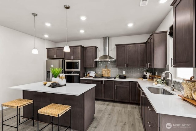 kitchen featuring dark brown cabinetry, wall chimney range hood, a sink, light wood finished floors, and appliances with stainless steel finishes