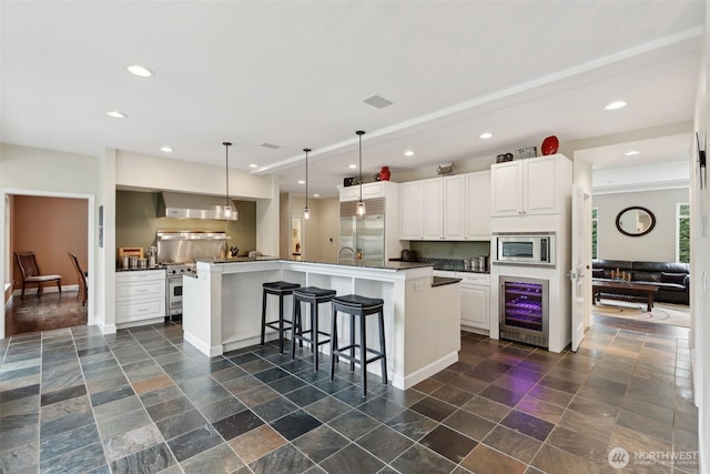 kitchen featuring built in appliances, dark countertops, beverage cooler, a center island with sink, and white cabinetry