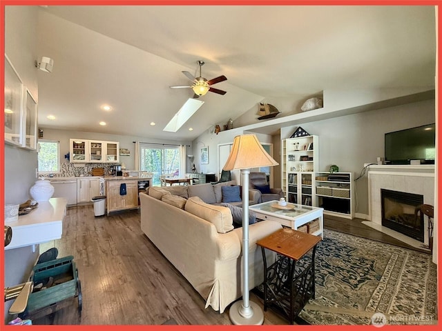 living area with a ceiling fan, dark wood-style flooring, a skylight, lofted ceiling, and a tiled fireplace