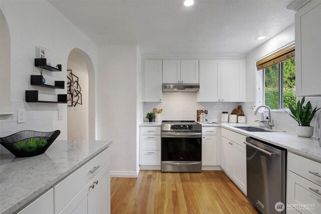 kitchen with a sink, arched walkways, stainless steel appliances, decorative backsplash, and under cabinet range hood