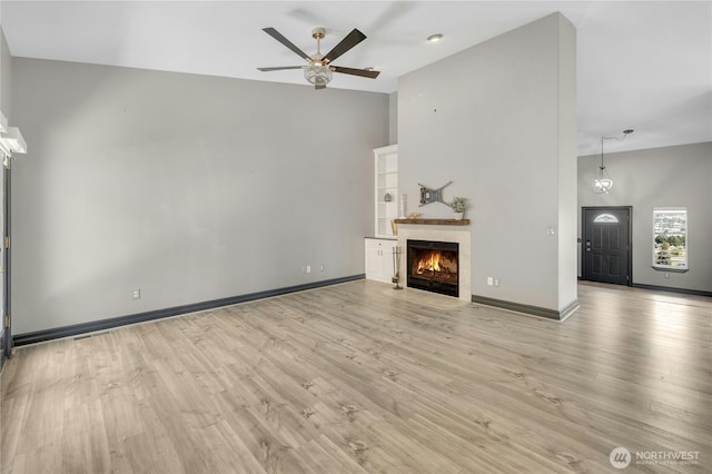 unfurnished living room featuring a warm lit fireplace, ceiling fan, light wood-style floors, high vaulted ceiling, and baseboards